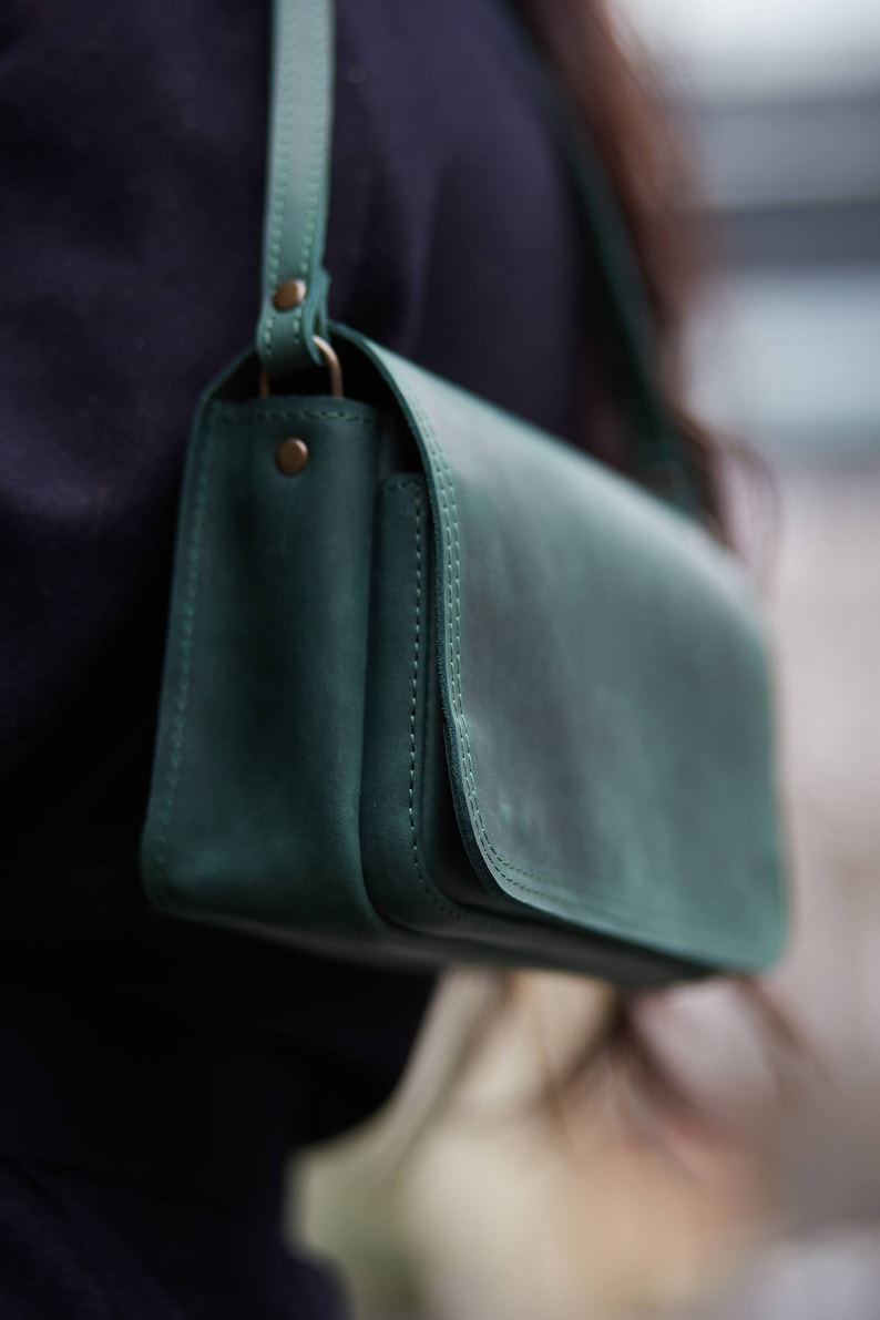 May include: A dark green leather shoulder bag with a flap closure and a single strap. The bag has a subtle distressed look and is made of high-quality leather.