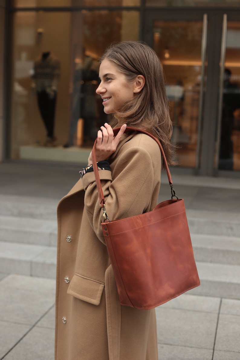 May include: A woman wearing a camel-colored coat carries a brown leather tote bag with a long strap over her shoulder.