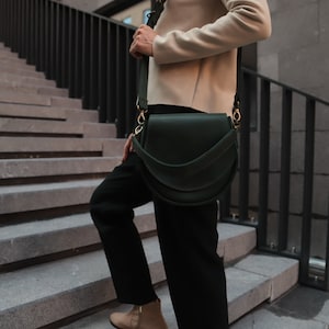 K&ouml;nnte beinhalten: Eine Person geht eine Treppe hinunter und tr&auml;gt eine dunkelgr&uuml;ne Lederschultertasche mit einer goldenen Schnalle.