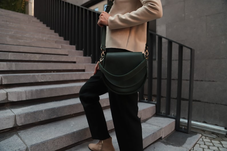 K&ouml;nnte beinhalten: Eine Person geht eine Treppe hinunter und tr&auml;gt eine dunkelgr&uuml;ne Lederschultertasche mit einer goldenen Schnalle.