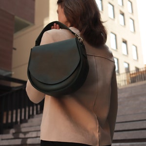 K&ouml;nnte beinhalten: Eine dunkelgr&uuml;ne Ledertasche mit einem Klappverschluss und einer silbernen Schnalle. Die Tasche wird &uuml;ber der Schulter einer Person getragen, die eine Treppe hinuntergeht.