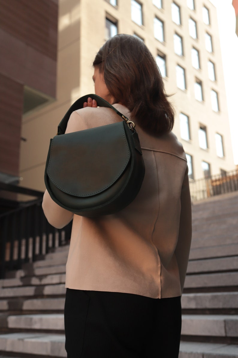 K&ouml;nnte beinhalten: Eine dunkelgr&uuml;ne Ledertasche mit einem Klappverschluss und einer silbernen Schnalle. Die Tasche wird &uuml;ber der Schulter einer Person getragen, die eine Treppe hinuntergeht.