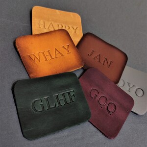 May include: Five leather patches with different names embossed on them. The names are "HAPPY", "WHAY", "JAN", "GLHIF", and "GOQ". The patches are arranged in a pile on a gray surface. Free embossing is offered.
