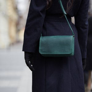 May include: A dark blue coat with a green leather shoulder bag. The bag has a flap closure and a strap.