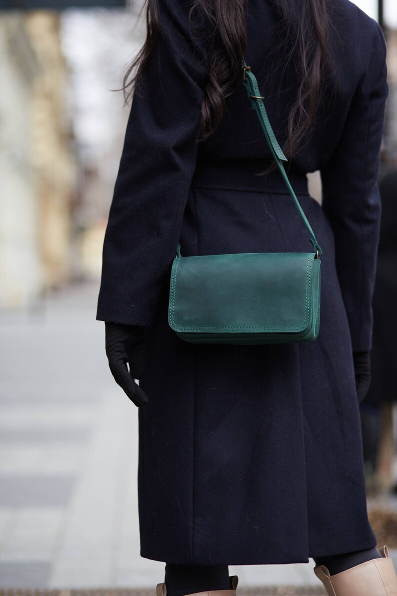 May include: A dark blue coat with a green leather shoulder bag. The bag has a flap closure and a strap.