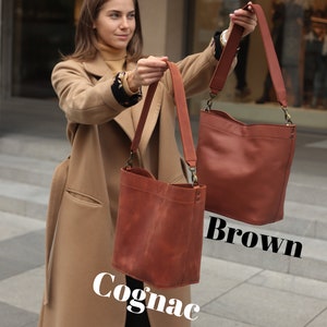 May include: Two leather tote bags, one brown and one cognac, held by a person wearing a camel coat. The bags have long straps and are made of a soft, supple leather.
