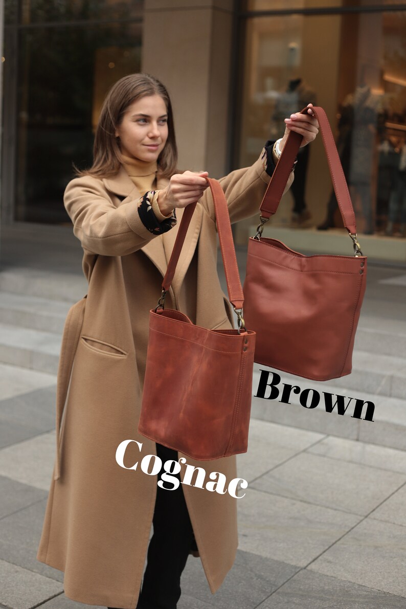 May include: Two leather tote bags, one brown and one cognac, held by a person wearing a camel coat. The bags have long straps and are made of a soft, supple leather.