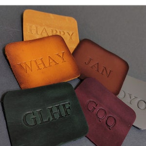 May include: Five leather patches with different colours and embossed text. The patches are brown, orange, maroon, green, and grey. The text on the patches reads "HAPPY", "WHAY", "JAN", "GLHF", and "GOQ".