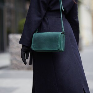 May include: A dark blue coat with a green leather crossbody bag. The bag has a long strap and a flap closure.