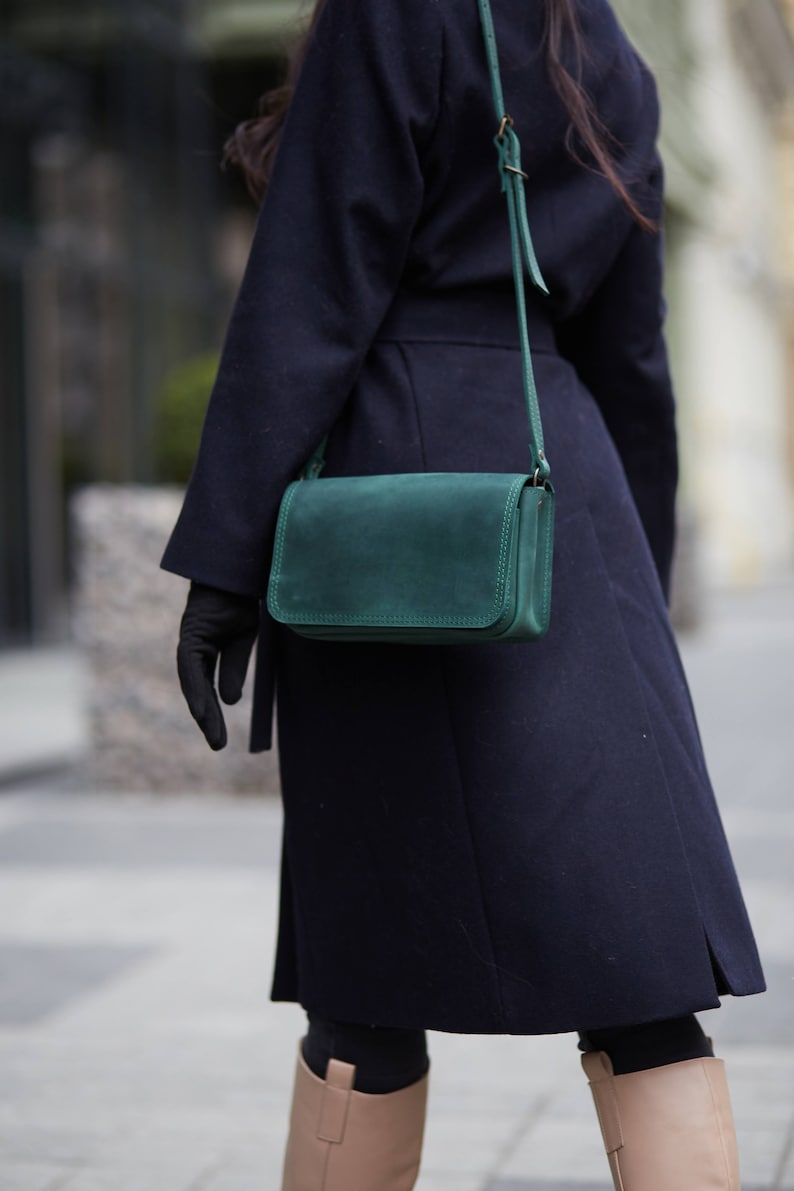 May include: A dark blue coat with a green leather crossbody bag. The bag has a long strap and a flap closure.