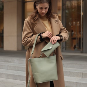 May include: A woman wearing a camel-colored coat carries a sage green leather tote bag with a smaller matching wallet.