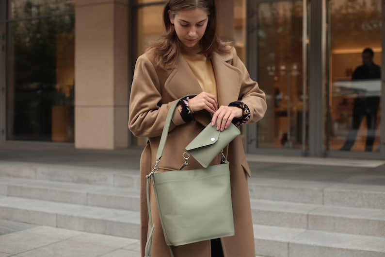 May include: A woman wearing a camel-colored coat carries a sage green leather tote bag with a smaller matching wallet.