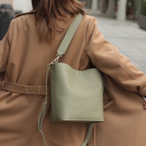 May include: A woman wearing a brown coat with a green leather bucket bag slung over her shoulder.