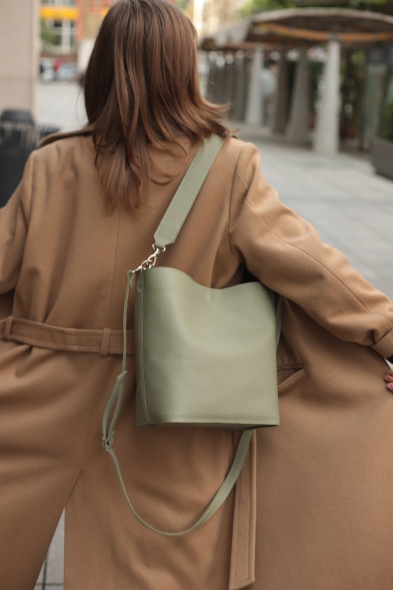 May include: A woman wearing a brown coat with a green leather bucket bag slung over her shoulder.