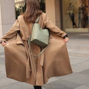 May include: A woman wearing a long brown coat with a green leather bucket bag slung over her shoulder. The coat has a belt and the bag has a long strap.
