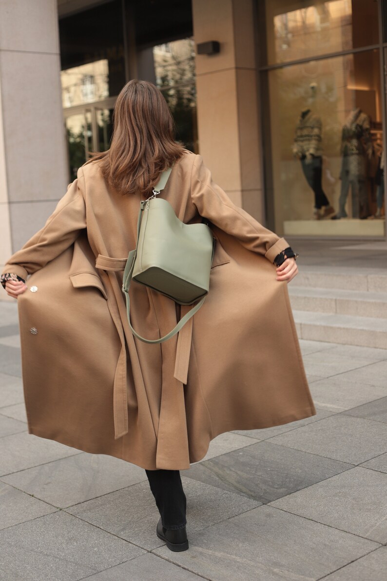 May include: A woman wearing a long brown coat with a green leather bucket bag slung over her shoulder. The coat has a belt and the bag has a long strap.