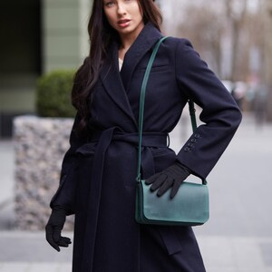 May include: A woman wearing a navy blue coat with a black beret and a green leather shoulder bag. She is wearing black gloves and has long dark hair.