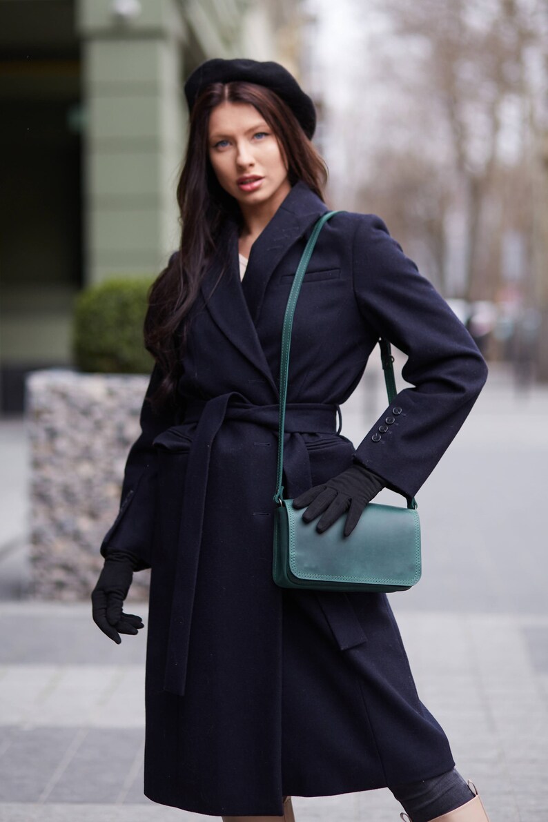 May include: A woman wearing a navy blue coat with a black beret and a green leather shoulder bag. She is wearing black gloves and has long dark hair.