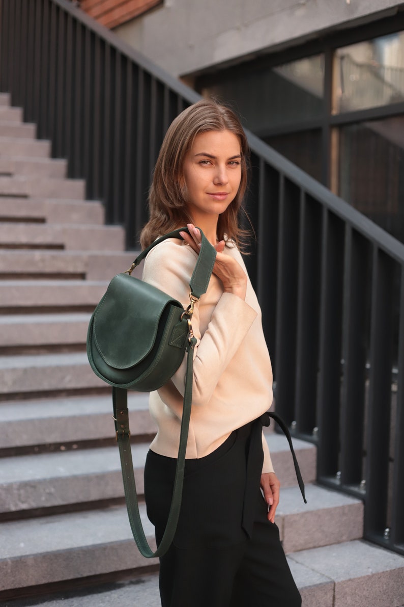 K&ouml;nnte beinhalten: Eine Frau in einem beigen Pullover und einer schwarzen Hose h&auml;lt eine dunkelgr&uuml;ne Ledertasche mit einem langen Riemen.
