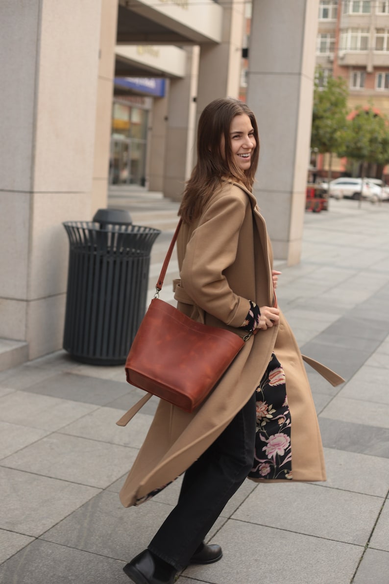 May include: A woman wearing a camel-colored coat with a floral lining and black pants. She is carrying a brown leather bucket bag with a long strap.