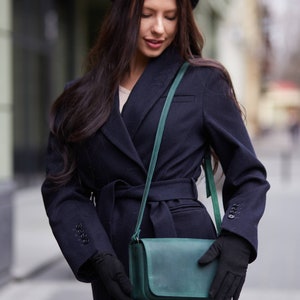 May include: A woman wearing a black coat with a belt, a black beret, and black gloves, holding a teal green leather crossbody bag.