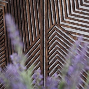 May include: A dark brown wooden wall panel featuring a series of vertical and angled slats that create a geometric pattern. The panel is composed of thin, parallel strips of wood with visible grain, arranged to form sharp, repeating chevron shapes.