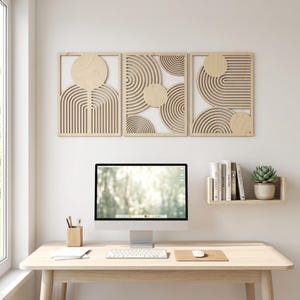 May include: Three rectangular light-colored wooden wall art panels featuring laser-cut geometric patterns of concentric circles and arches. The panels are arranged horizontally above a computer monitor on a desk with a small shelf holding books and a plant.