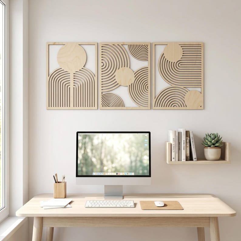 May include: Three rectangular light-colored wooden wall art panels featuring laser-cut geometric patterns of concentric circles and arches. The panels are arranged horizontally above a computer monitor on a desk with a small shelf holding books and a plant.