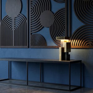 May include: A geometric table lamp featuring stacked rectangular blocks in brushed brass and matte black finishes, emitting a warm glow from its center. The lamp sits on a thin, black metal console table against a blue wall with dark wood panels.