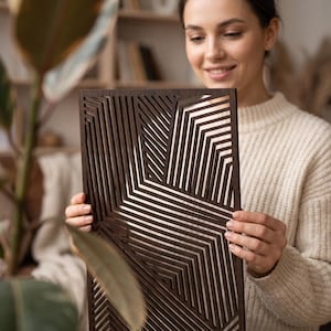 May include: A rectangular dark brown wooden wall panel featuring a laser-cut geometric pattern of parallel lines that create angular, intersecting shapes. The panel has a smooth finish and a series of open slots throughout the entire design.