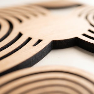 May include: A light brown wooden object featuring intricate laser-cut concentric circular patterns. The material has a natural wood grain texture with dark, charred edges along the cut lines, creating a high-contrast geometric design.