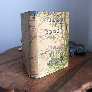 May include: A vintage hardback book titled "Cider with Rosie" by Laurie Lee. The book cover features a green and brown illustration of a countryside landscape with a house and trees.