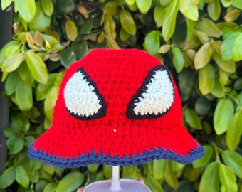 May include: Red crocheted bucket hat with a black and white spider-man design. The hat has a navy blue brim.