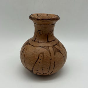 May include: A small, brown ceramic vase with a rounded body and a narrow neck. The vase is adorned with dark brown, abstract patterns. The design includes curved lines and dotted accents, creating a unique, handcrafted appearance. The vase has a textured surface.