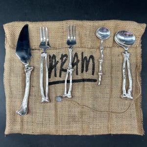 May include: A five-piece silver-toned flatware set featuring handles shaped like human bones, arranged on a tan burlap fabric pouch with the word ARAM printed in black ink. The set includes a knife, two forks, and two spoons of varying sizes.