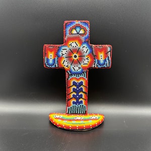 May include: A colorful beaded cross with a red, blue, and yellow floral design. The cross is on a stand with a similar beaded design.