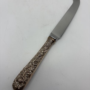 May include: A cheese knife featuring a polished silver-tone metal blade with a forked tip and a decorative handle crafted from tarnished silver-tone metal, displaying an intricate embossed floral and leaf pattern throughout the entire handle surface.