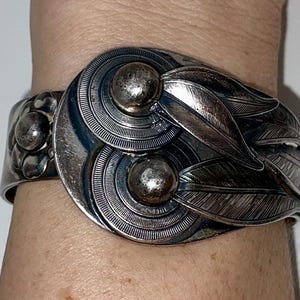 May include: A silver-toned cuff bracelet with an ornate design. The bracelet features two large, circular elements with concentric circles and two spherical accents. Leaf-shaped details and floral accents complete the design.