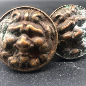 May include: Two antique bronze-colored cabinet knobs with a lion's head design. The knobs are round with a detailed relief of a lion's face, showing signs of age and wear. The metal has a patina with hints of green.