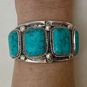May include: A silver bracelet with four turquoise stones set in a square shape. The bracelet has a detailed design with a rope-like border and a floral pattern.