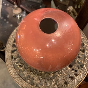 May include: A pink glass globe sits atop a decorative gold metal stand with intricate floral designs. The globe has a small opening at the top.