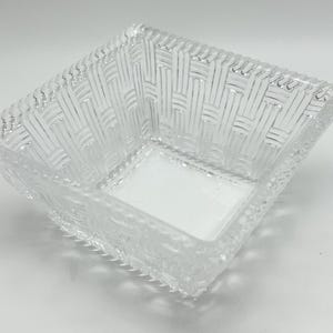 Puede incluir: Cuenco cuadrado de vidrio transparente con diseño de cesta tejida. El cuenco tiene un patrón texturizado y un borde decorativo. El cuenco está vacío y se encuentra sobre una superficie blanca.