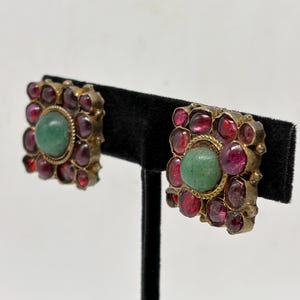 May include: A pair of square-shaped earrings featuring a central round green stone surrounded by multiple smaller, deep red, cabochon-cut stones set in a gold-toned metal frame. The design includes a textured, rope-like border around the center.