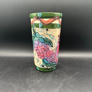 May include: A green ceramic mug with a hand-painted design of grapes and vines. The grapes are pink, purple, and green. The mug has a white background with green and brown stripes.