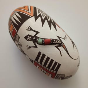 May include: A white ceramic pot with a black, red, and orange geometric design. A black and red lizard with a turquoise stripe is painted on the pot.