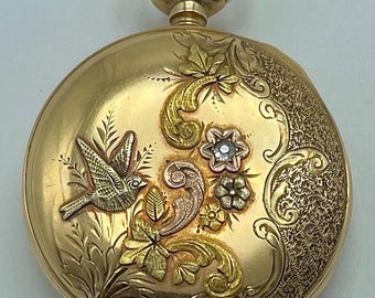 May include: A gold pocket watch with intricate floral and bird engravings. The watch features a detailed design with a flying bird, flowers, and ornate patterns. The watch has a loop at the top for a chain.