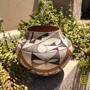 May include: A round ceramic pot featuring a cream-colored base with intricate hand-painted geometric patterns in black and terracotta brown. The vessel has a wide, open mouth and a smooth, matte finish, showcasing detailed line work and shapes.