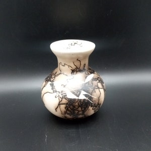 May include: A small, off-white ceramic vase with a rounded body and a narrow neck. The vase is decorated with intricate black lightning-like patterns and a few white accents. The overall design is organic and artistic.