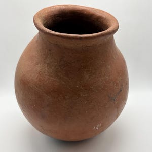 May include: A round, terracotta-colored clay pot with a wide, open mouth and a thick, rounded rim. The surface of the vessel has a matte, textured finish with natural variations in color, showing a simple, bulbous shape and a deep, dark interior.