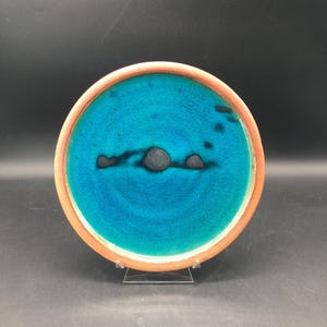 May include: A round ceramic plate with a turquoise center and a brown rim. The turquoise surface has a crackle glaze effect and features a dark, abstract design. The plate is displayed on a clear stand.